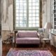 Une pièce cosy avec un canapé lavande, deux tables d'appoint, une lampe et un tapis coloré près de grandes fenêtres vintage respirant la qualité parisienne. Canape Paris by Atelier Bis