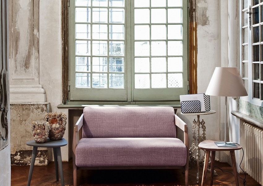 Une pièce cosy avec un canapé lavande, deux tables d'appoint, une lampe et un tapis coloré près de grandes fenêtres vintage respirant la qualité parisienne. Canape Paris by Atelier Bis