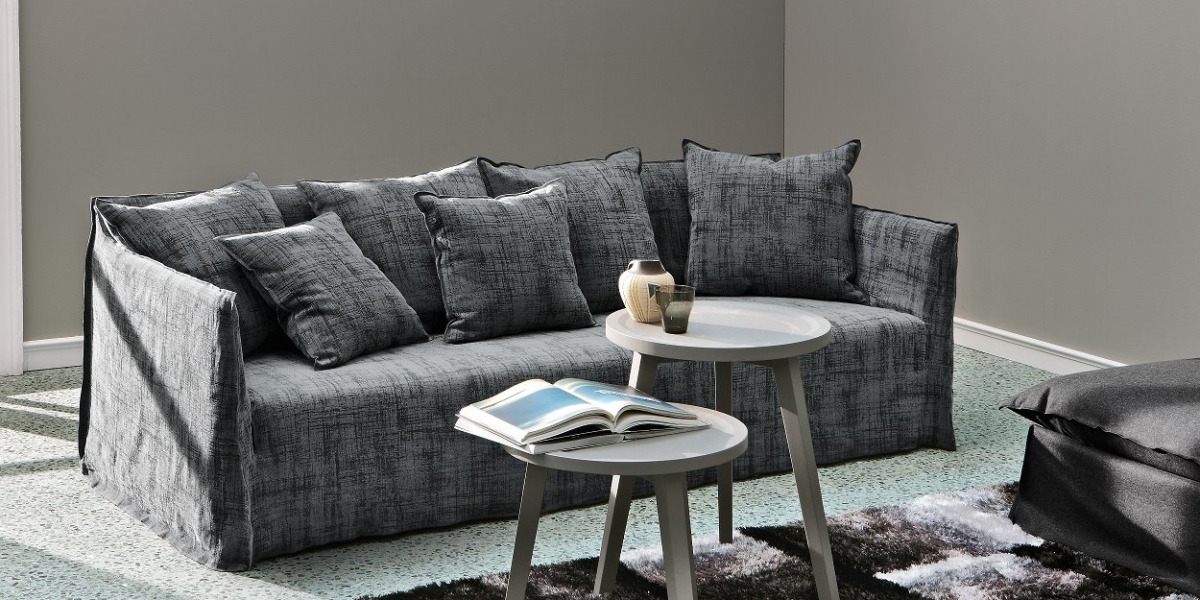 Canapé gris avec plusieurs coussins, deux tables d'appoint rondes blanches avec des livres et des décorations, et murs gris dans un showroom moderne rappelant l'élégance parisienne. Canape Paris by Atelier Bis
