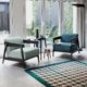 Deux fauteuils modernes se font face près d'une petite table dans un showroom parisien lumineux, baigné de lumière traversant de grandes fenêtres et complété par un tapis géométrique chic. Canape Paris by Atelier Bis