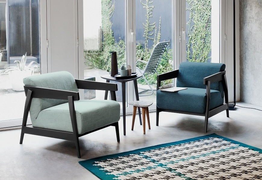 Deux fauteuils modernes se font face près d'une petite table dans un showroom parisien lumineux, baigné de lumière traversant de grandes fenêtres et complété par un tapis géométrique chic. Canape Paris by Atelier Bis