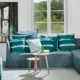 Salon parisien moderne avec un canapé bleu canard, des coussins rayés et une table ronde en bois. Une grande fenêtre met en valeur une végétation luxuriante, ajoutant une touche de qualité naturelle à cet espace chic. Canape Paris by Atelier Bis