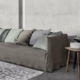 Un canapé gris texturé orné de plusieurs coussins gris et verts trône élégamment à côté d'une petite table d'appoint sur un sol gris chic, rappelant un showroom parisien élégant. Canape Paris by Atelier Bis