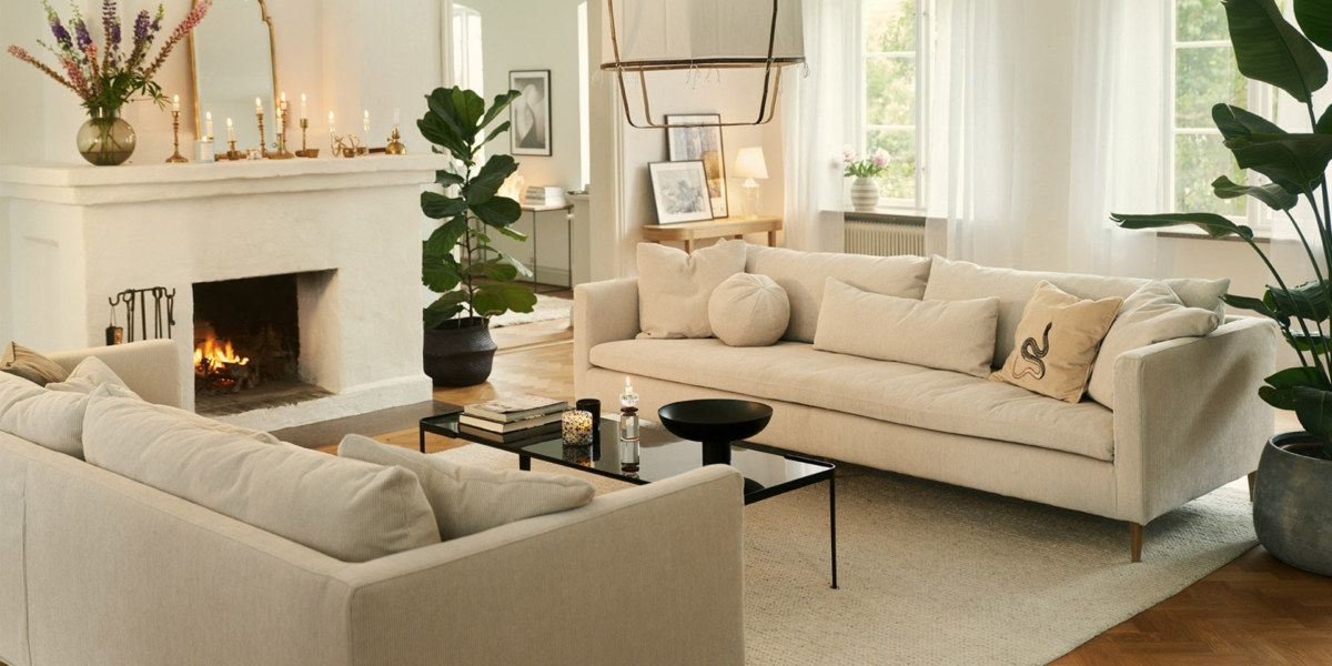 Salon chaleureux avec canapés beiges, plantes luxuriantes, cheminée chaleureuse et bougies élégamment placées sur le manteau. De grandes fenêtres de style showroom inondent l'espace de lumière naturelle. Canape Paris by Atelier Bis