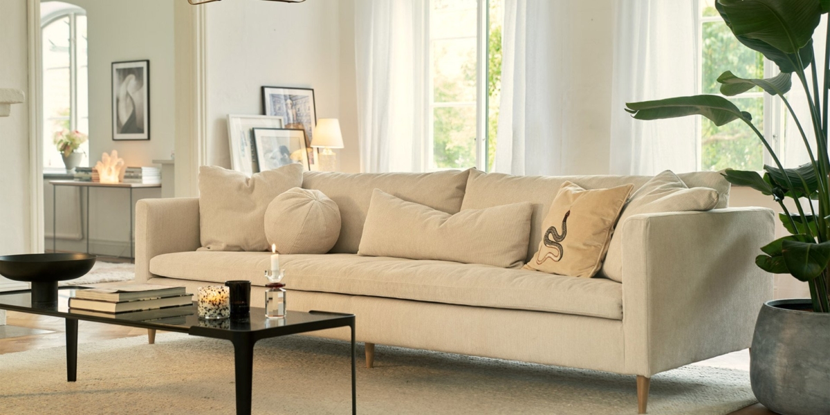 Un canapé beige avec coussins orne le salon lumineux, où une table noire ornée de bougies et de livres ajoute du charme. Une grande plante complète ce décor d'inspiration parisienne, offrant une touche d'élégance et de qualité. Canape Paris by Atelier Bis