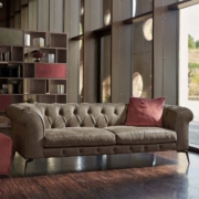 Un salon moderne dispose d'un canapé en cuir orné de coussins rouges, d'un repose-pieds, de grandes fenêtres et d'une bibliothèque en bois, le tout reflétant un équilibre exquis entre prix et qualité. Canape Paris by Atelier Bis
