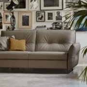 Une pièce chaleureuse est dotée d'un canapé beige orné d'un coussin marron, de photos encadrées et d'une grande plante, créant une ambiance rappelant une salle d'exposition élégante. Canape Paris by Atelier Bis
