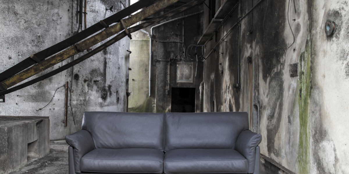 Un canapé gris foncé dans un décor industriel patiné avec des poutres métalliques et des murs en béton texturé, rappelant un showroom parisien chic. Canape Paris by Atelier Bis