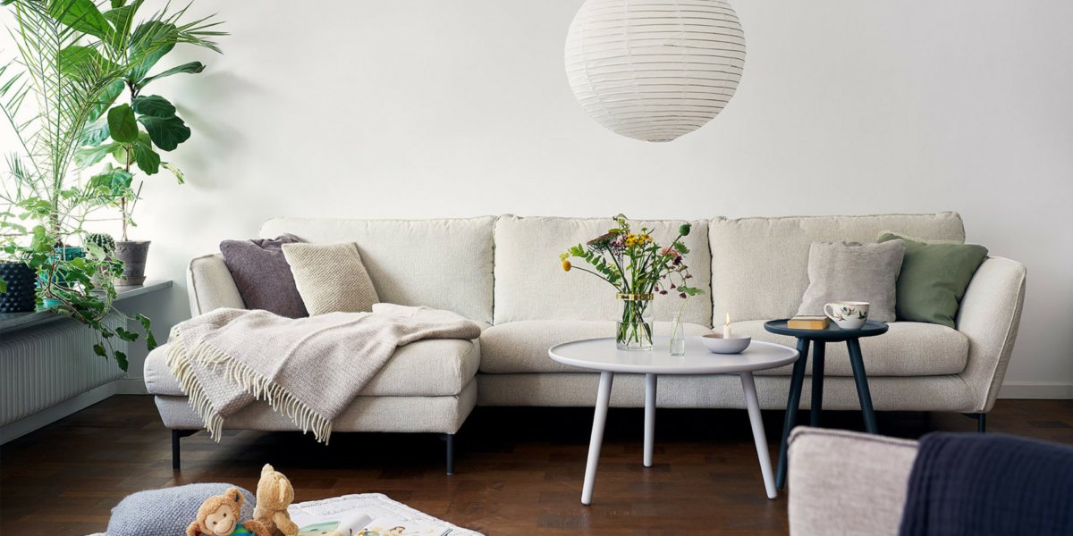 Un salon parisien cosy doté d'un canapé blanc, de plantes luxuriantes, de tables basses élégantes et d'une lampe suspendue chic qui donne l'impression d'entrer dans un showroom accueillant. Canape Paris by Atelier Bis