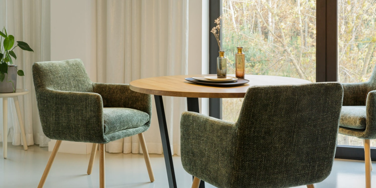 La salle à manger comprend une table ronde en bois, des chaises rembourrées vertes et une plante en pot près de grandes fenêtres avec des rideaux, rappelant une salle d'exposition où chaque détail témoigne de l'équilibre parfait entre prix et qualité. Canape Paris by Atelier Bis