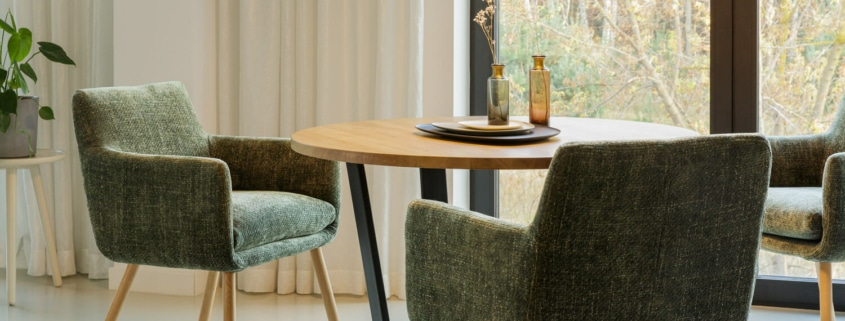 La salle à manger comprend une table ronde en bois, des chaises rembourrées vertes et une plante en pot près de grandes fenêtres avec des rideaux, rappelant une salle d'exposition où chaque détail témoigne de l'équilibre parfait entre prix et qualité. Canape Paris by Atelier Bis