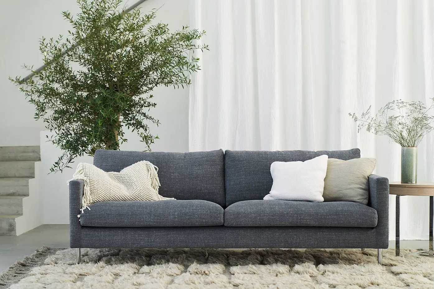 Un canapé gris moderne avec des coussins blancs, une couverture douillette et une grande plante dans un salon lumineux. Canape Paris by Atelier Bis