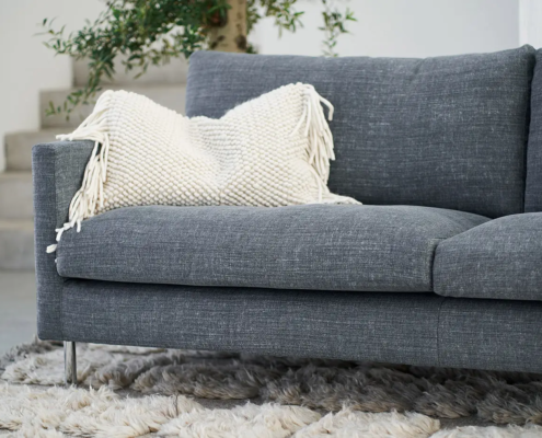 Un canapé gris avec un coussin à franges blanches sur un tapis à poils longs texturé, à côté d'une plante. Canape Paris by Atelier Bis
