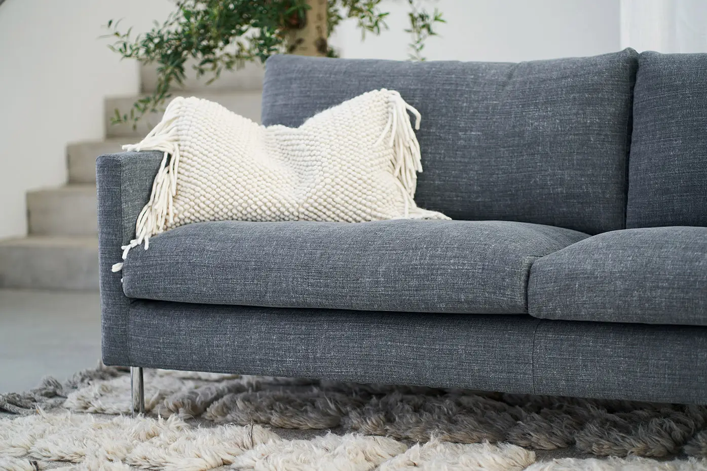 Un canapé gris avec un coussin à franges blanches sur un tapis à poils longs texturé, à côté d'une plante. Canape Paris by Atelier Bis
