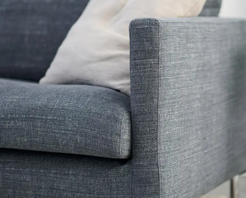 Un canapé en tissu gris avec un coussin léger repose sur un tapis beige à poils longs. Canape Paris by Atelier Bis