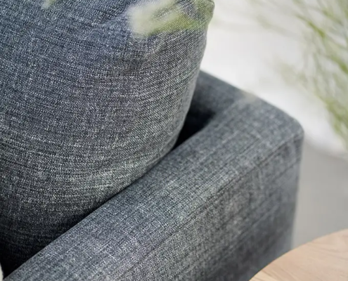 Gros plan d'un canapé en tissu gris avec des coussins moelleux à côté d'une table en bois clair et d'un feuillage vert. Canape Paris by Atelier Bis