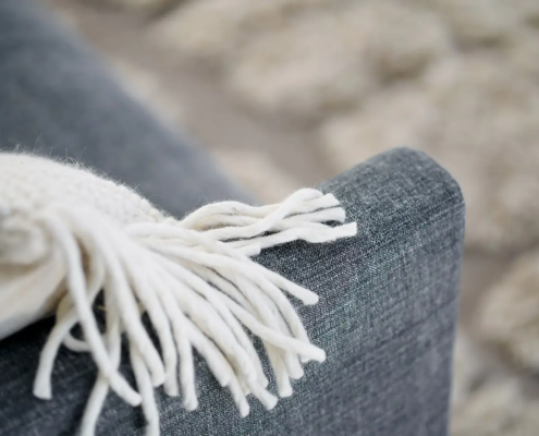 Gros plan d'un canapé gris avec un jeté en tricot blanc drapé sur l'accoudoir, tapis flou en arrière-plan. Canape Paris by Atelier Bis