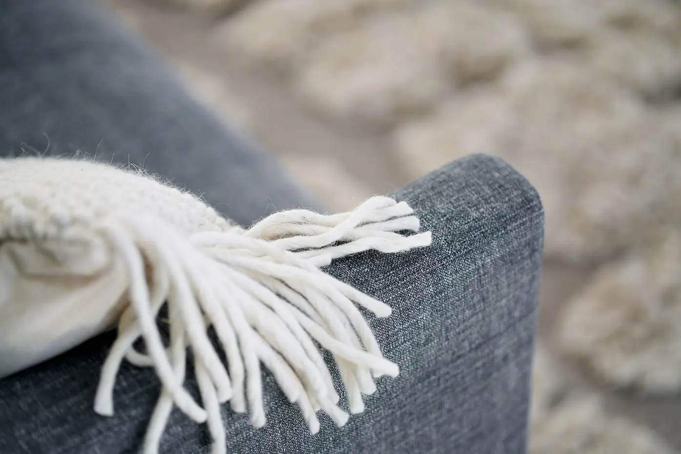 Gros plan d'un canapé gris avec un jeté en tricot blanc drapé sur l'accoudoir, tapis flou en arrière-plan. Canape Paris by Atelier Bis
