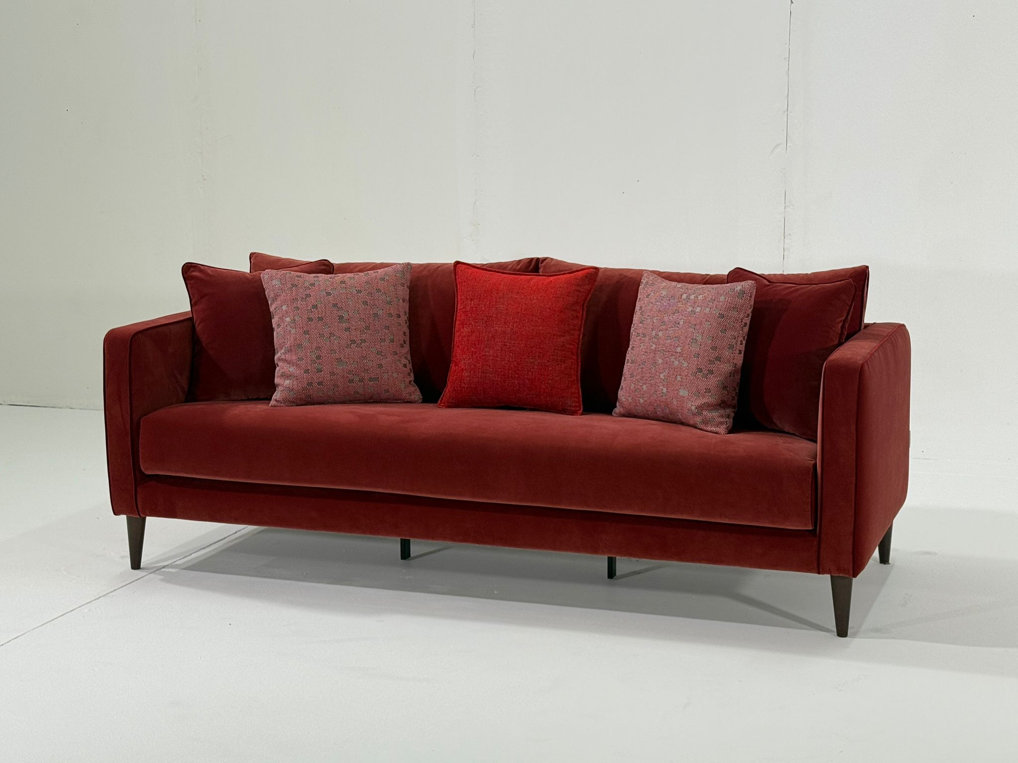 Canapé en velours rouge avec cinq coussins assortis à motifs et unis, disposés sur un fond blanc uni. Canape Paris by Atelier Bis