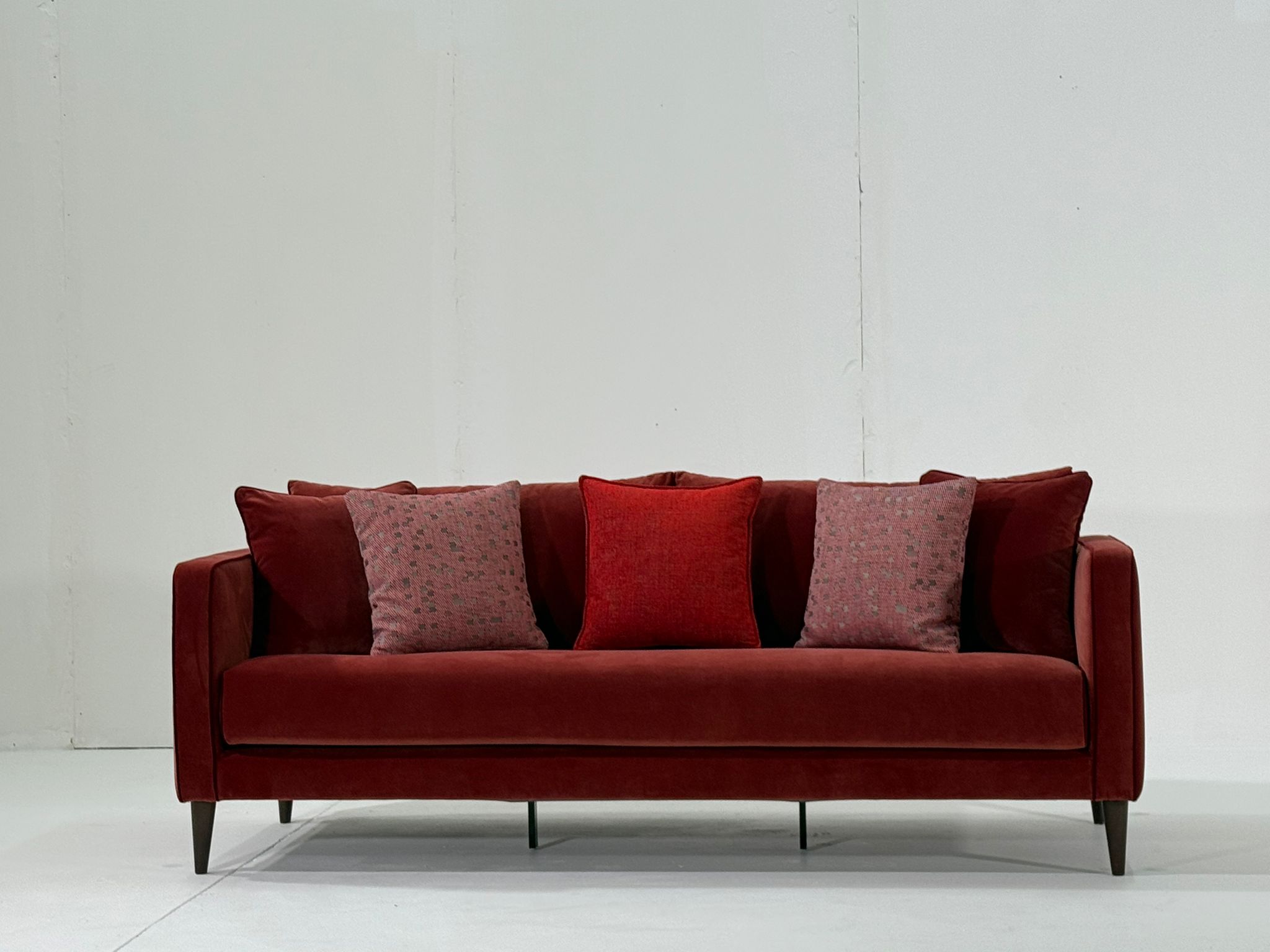Un canapé rouge avec cinq coussins, posé contre un mur blanc uni et un sol blanc. Canape Paris by Atelier Bis