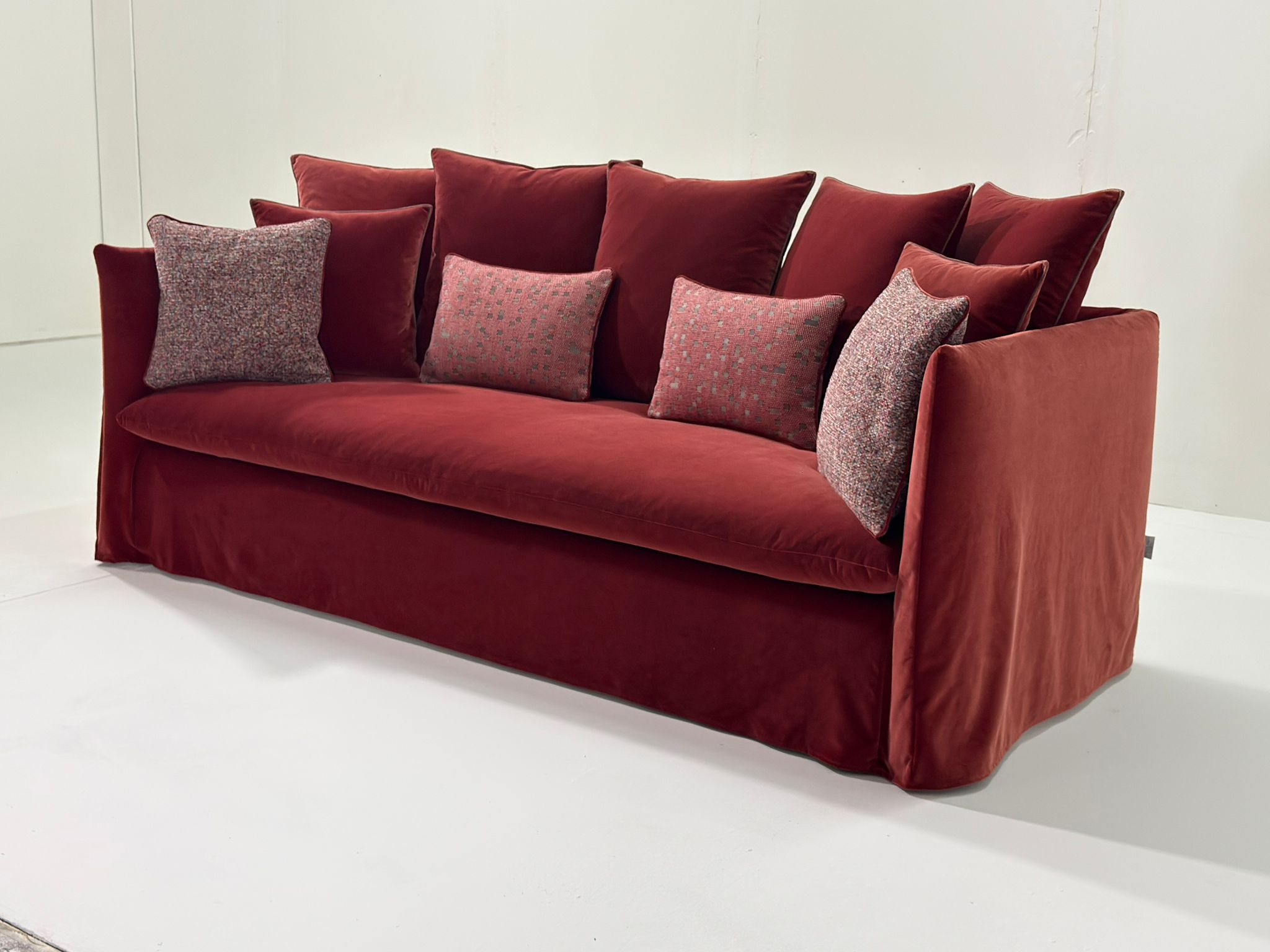 Un canapé moderne en velours rouge avec six coussins à motifs dans une pièce blanche minimaliste. Canape Paris by Atelier Bis