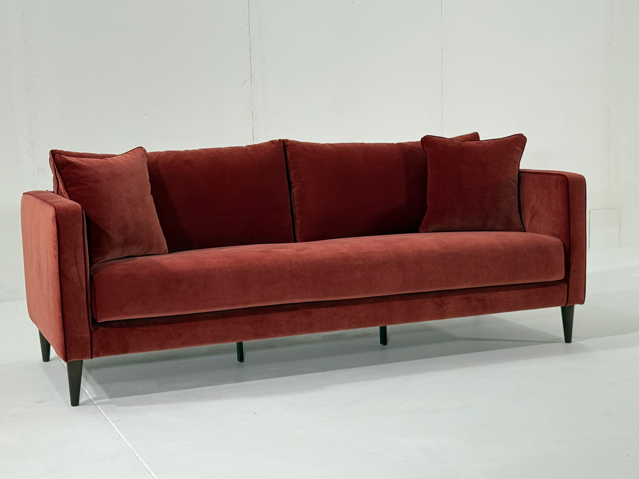 Un canapé moderne en velours rouge avec deux coussins assortis contre un mur blanc uni. Canape Paris by Atelier Bis