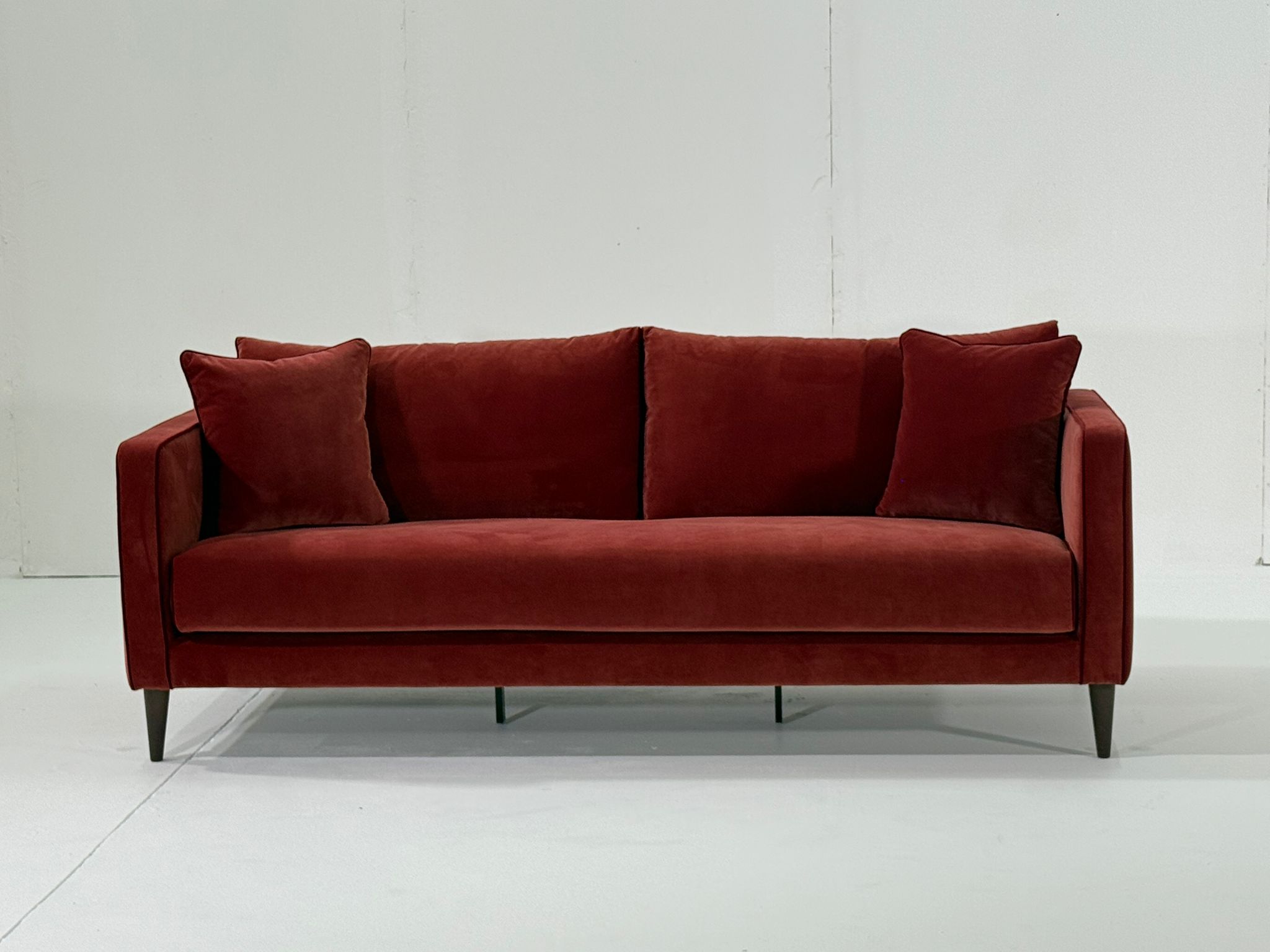 Un canapé moderne en velours rouge avec deux coussins assortis, sur un fond blanc uni. Canape Paris by Atelier Bis