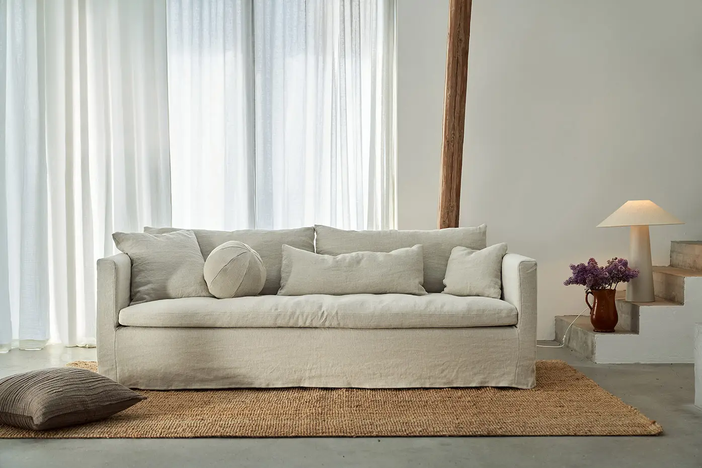 Un canapé beige avec des coussins repose sur un tapis tressé dans un salon lumineux et minimaliste avec des rideaux transparents. Canape Paris by Atelier Bis