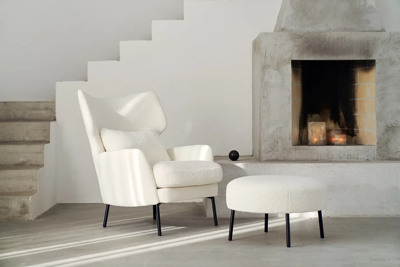 Fauteuil et ottoman blancs modernes à côté d'une cheminée en béton et d'un escalier minimaliste dans une pièce lumineuse. Canape Paris by Atelier Bis