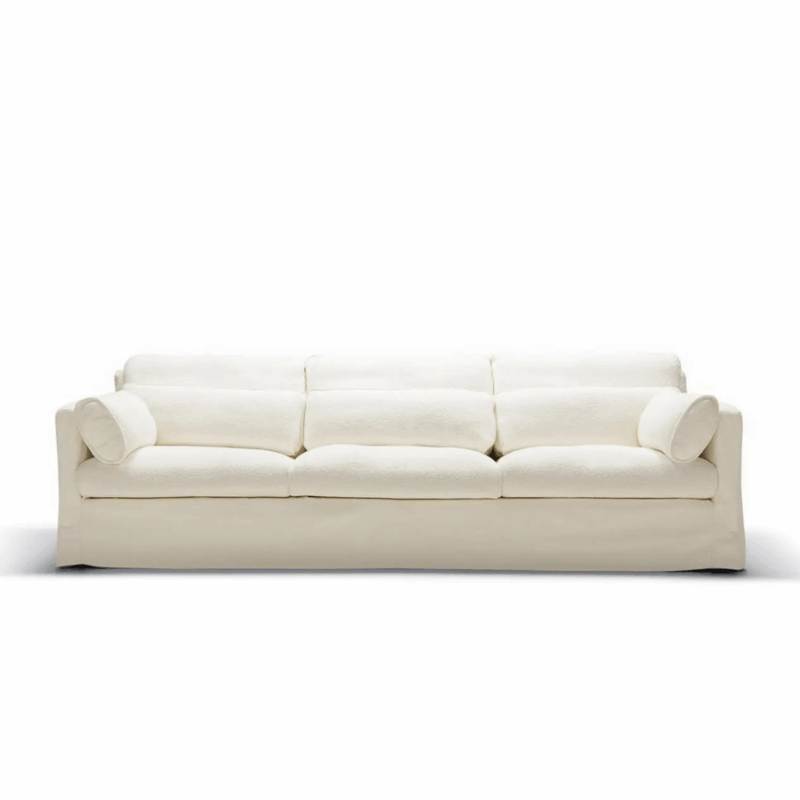 Canapé blanc à trois places avec des coussins en peluche et deux oreillers cylindriques, sur un fond blanc uni. Canape Paris by Atelier Bis
