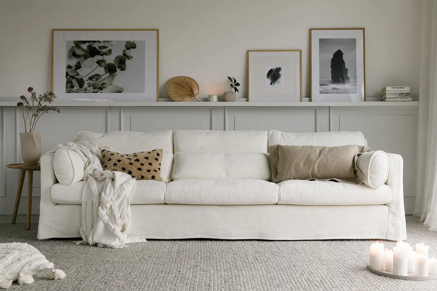 Un canapé blanc confortable avec des coussins neutres et une couverture, dans un salon minimaliste avec des œuvres d'art encadrées et des bougies. Canape Paris by Atelier Bis