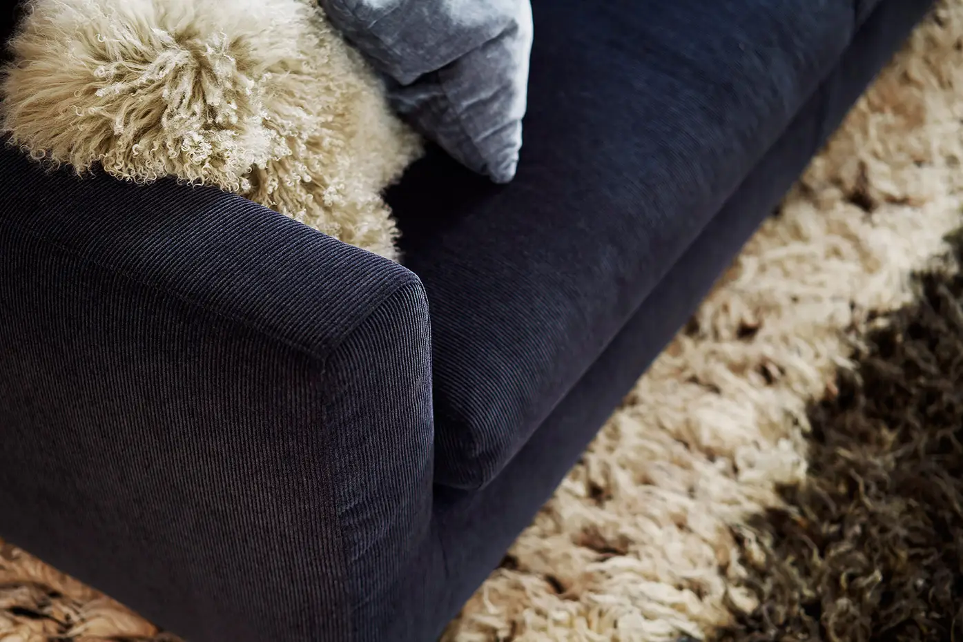 Gros plan sur un canapé bleu foncé avec un jeté et un coussin beige duveteux à côté d'un tapis beige hirsute. Canape Paris by Atelier Bis